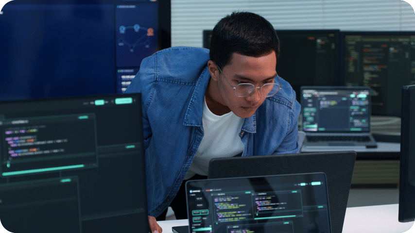 A man in glasses and a denim shirt leans over a desk studying code on multiple monitors in a dark office.