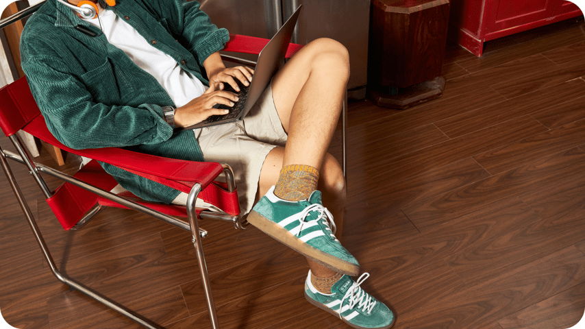 Person typing on a laptop in a red chair, wearing a green corduroy jacket, beige shorts, mustard socks, and green suede Adidas Gazelle trainers.