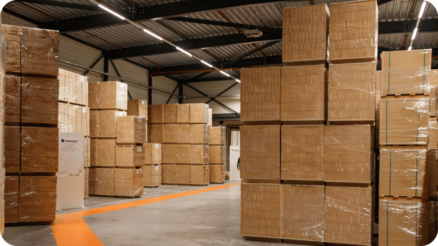 Warehouse interior with stacked cardboard boxes wrapped in plastic, an orange floor path, and overhead strip lighting.