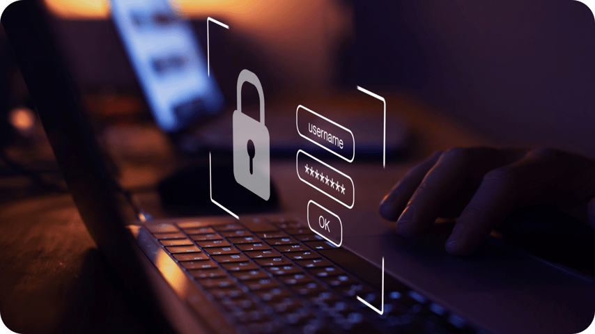 A hand on a laptop at night, with a glowing padlock icon and a holographic username and password login overlay symbolising cybersecurity.