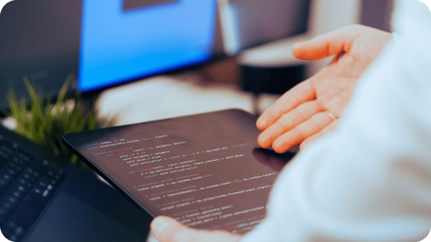 Close-up of a person holding a tablet displaying lines of code, with a laptop keyboard and monitor visible in the background.