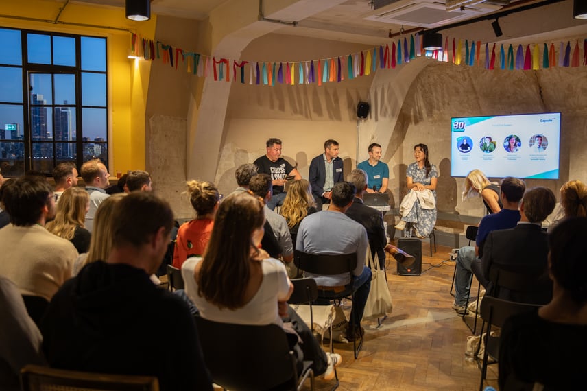 Panel discussion at Capsule’s B-Corp 30 event in a modern venue with wooden floors and a yellow wall. Four speakers are seated at the front, with a large screen displaying presentation slides featuring profile images and text.