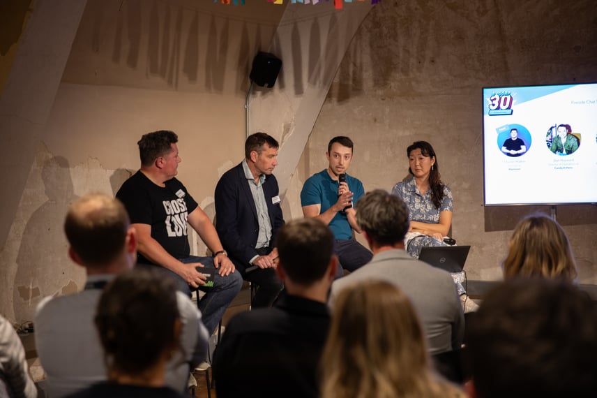 Panel discussion at Capsule’s B-Corp 30 event. Four speakers are seated at the front of the room, with one person holding a microphone while speaking. A large screen to the right displays the event branding “B-Corp 30” and profile images of two individuals.