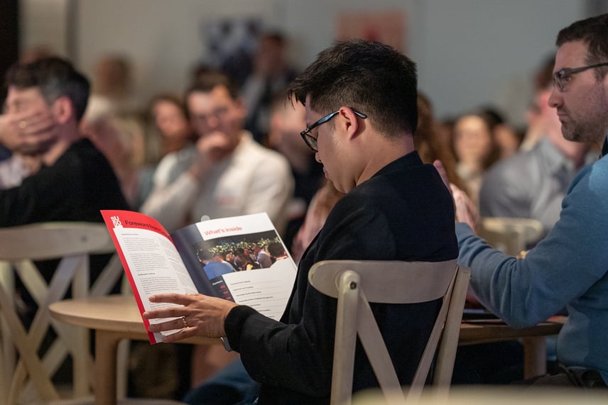 An attendee reading the ScaleUp50 programme booklet during the event, with a full audience visible and engaged in the background.