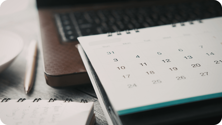 A desk calendar, notebook and pen on a wooden desk beside a laptop