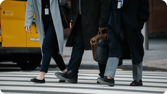Three people wearing long coats and carrying bags cross a city street at a marked pedestrian crossing, with a yellow vehicle visible in the background.