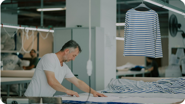 Tailor working with striped fabric at a cutting table in a garment factory, with a completed striped top displayed on a hanger