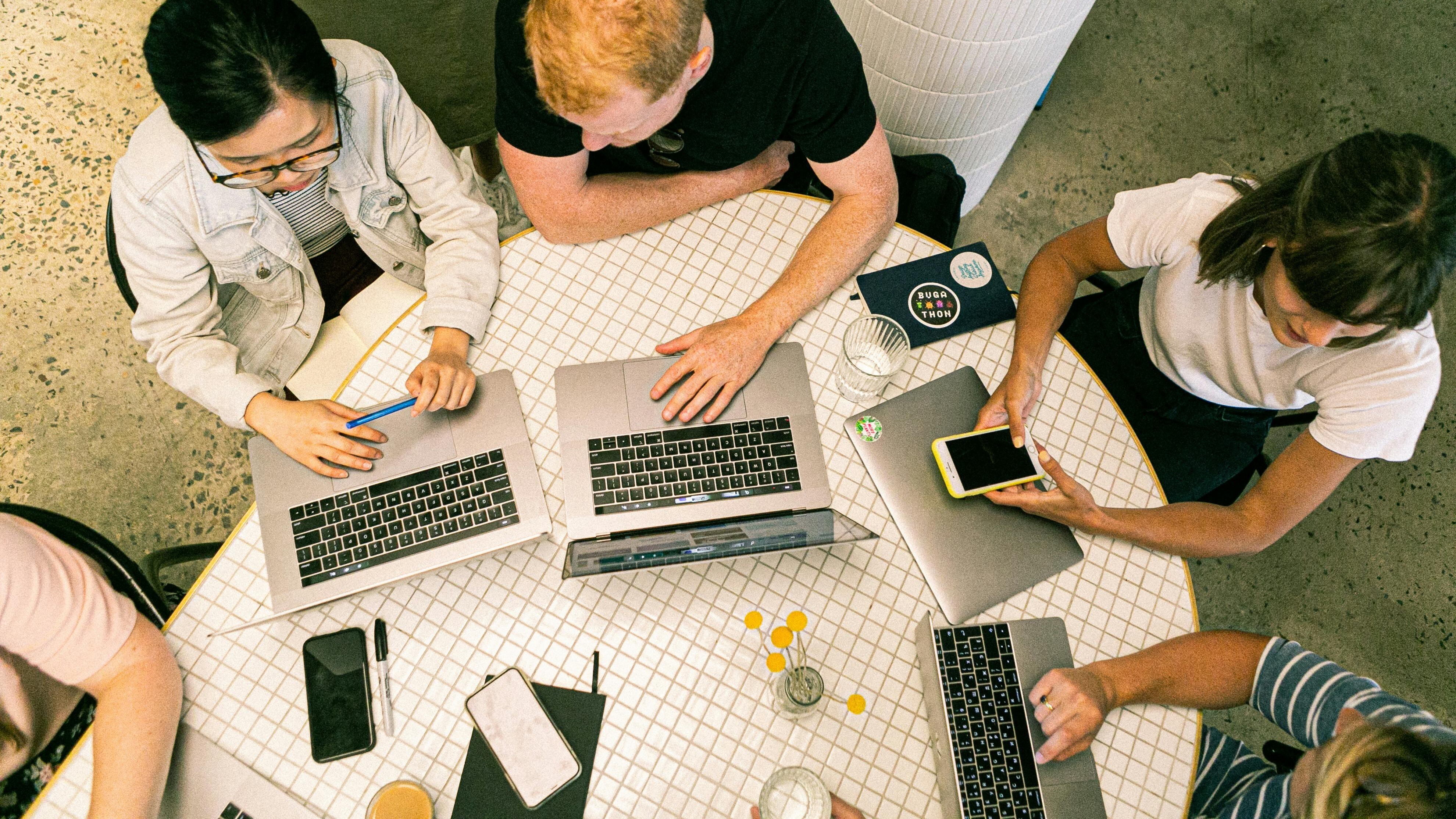 Top-down view of a round white-tiled table with several open laptops, a smartphone, notebooks, and pens. Multiple people are seated around the table, actively working and collaborating. 