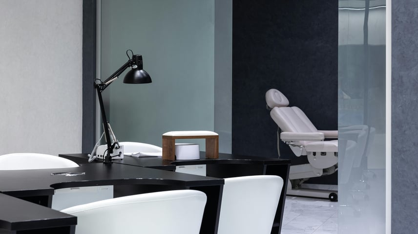 Modern, minimalist office or salon space with black desks and white chairs in the foreground, a black adjustable desk lamp, and a manicure station setup. In the background, there is a white reclining treatment chair on wheels, positioned against dark and light-colored walls