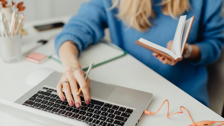 Person wearing a blue sweater sitting at a white desk, using a laptop with one hand while holding an open notebook in the other. The desk has scattered items including a pencil, a charging cable, and stationery.