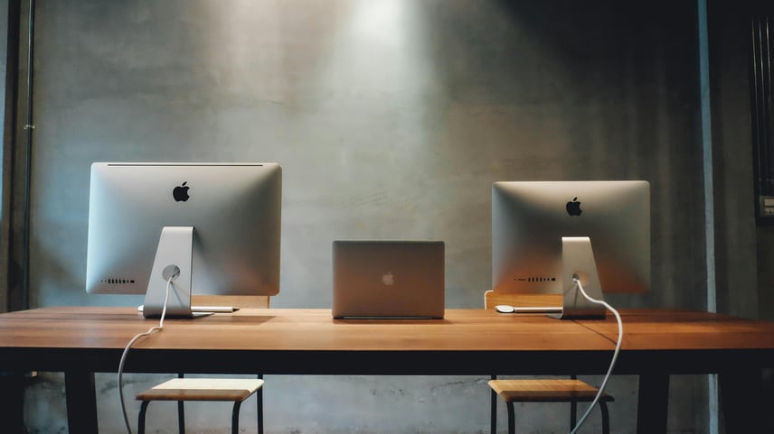 A minimalist workspace featuring a wooden table with two large Apple desktop computers and a closed Apple laptop placed between them. Each desktop has a white power cable connected, and two simple wooden stools are positioned in front of the table. The background consists of a plain gray wall with soft overhead lighting creating a focused, modern aesthetic.